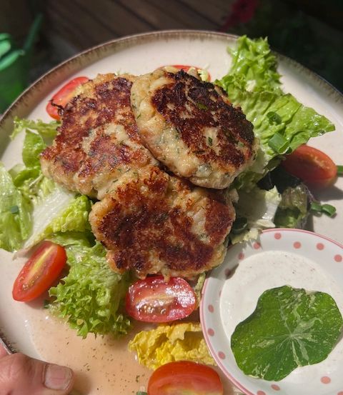 Ein Teller mit drei gebratenen Patties auf einem Bett aus Salat, begleitet von einer kleinen Schüssel grüner Sauce und geschnittenen Tomaten.