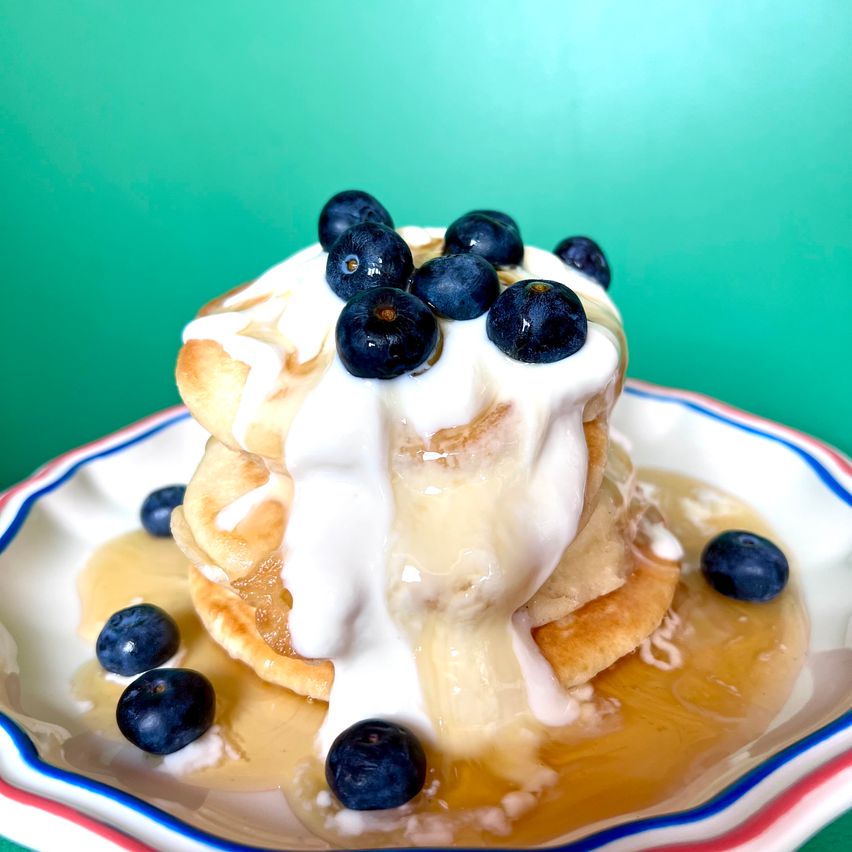A plate of pancakes topped with whipped cream, blueberries, and drizzled maple syrup on a green background.