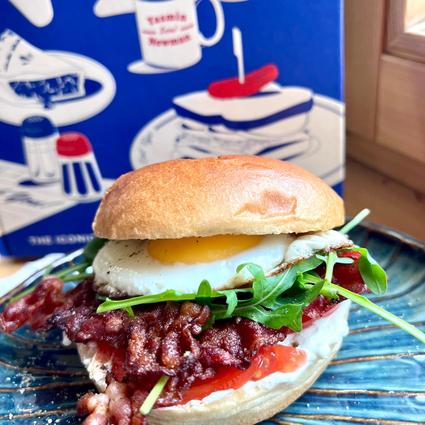 A sandwich with bacon, a fried egg, and arugula on a blue plate, with a box of 'Essemble' soup in the background.
