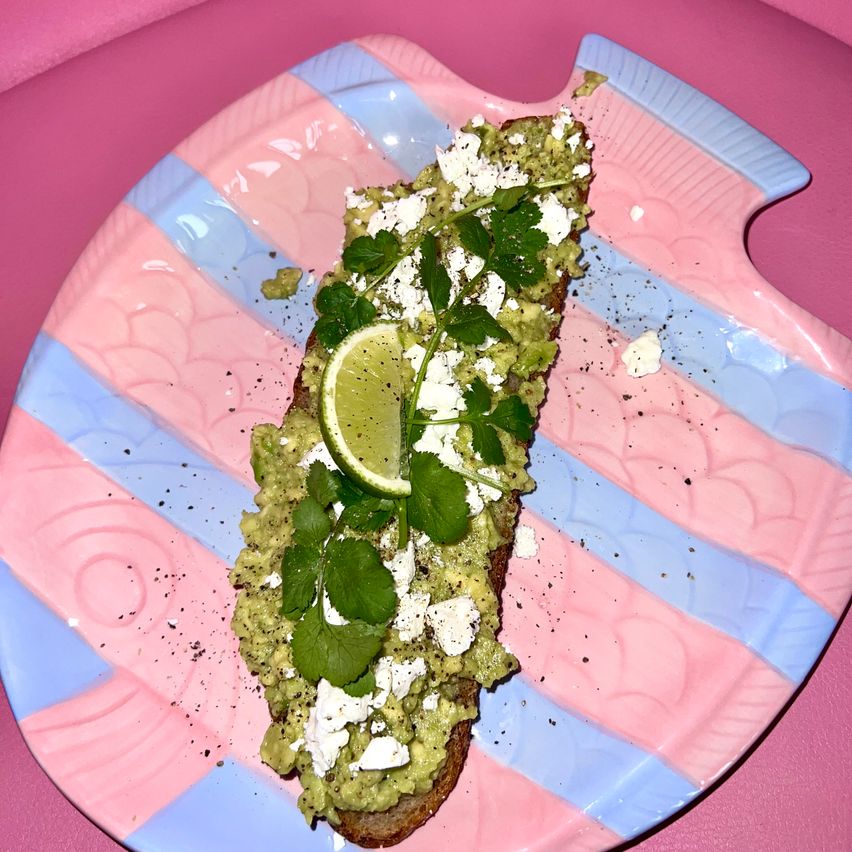 A toasted bread with guacamole, feta cheese, cilantro, and a lime wedge on a pink and blue striped plate.