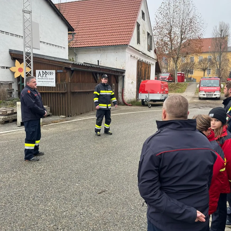 Eine Gruppe von Menschen versammelt sich im Freien. Zwei Männer in Uniform stehen auf der Straße. Dahinter ist ein Feuerwehrfahrzeug geparkt. Gebäude und ein Schild mit einer Blume sind sichtbar.
