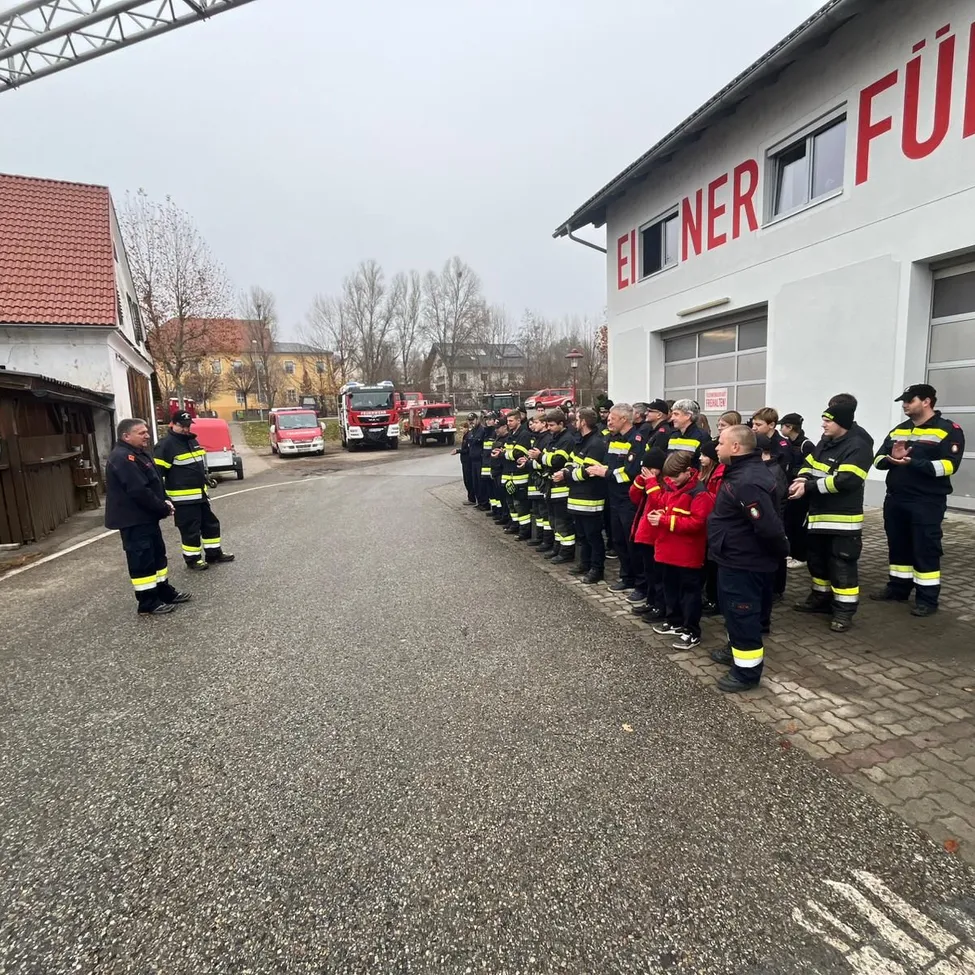 Eine Gruppe von Feuerwehrleuten steht in einer Reihe vor einer Feuerwache, mit einem roten LKW im Hintergrund.