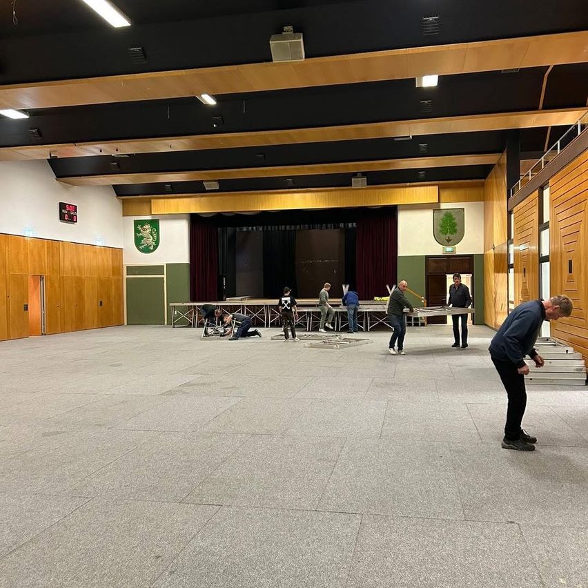 Mehrere Personen arbeiten in einem großen, leeren Raum. Holzbalken stützen die Decke, und eine Bühne ist am anderen Ende sichtbar. Holzwände und Türen sind vorhanden.