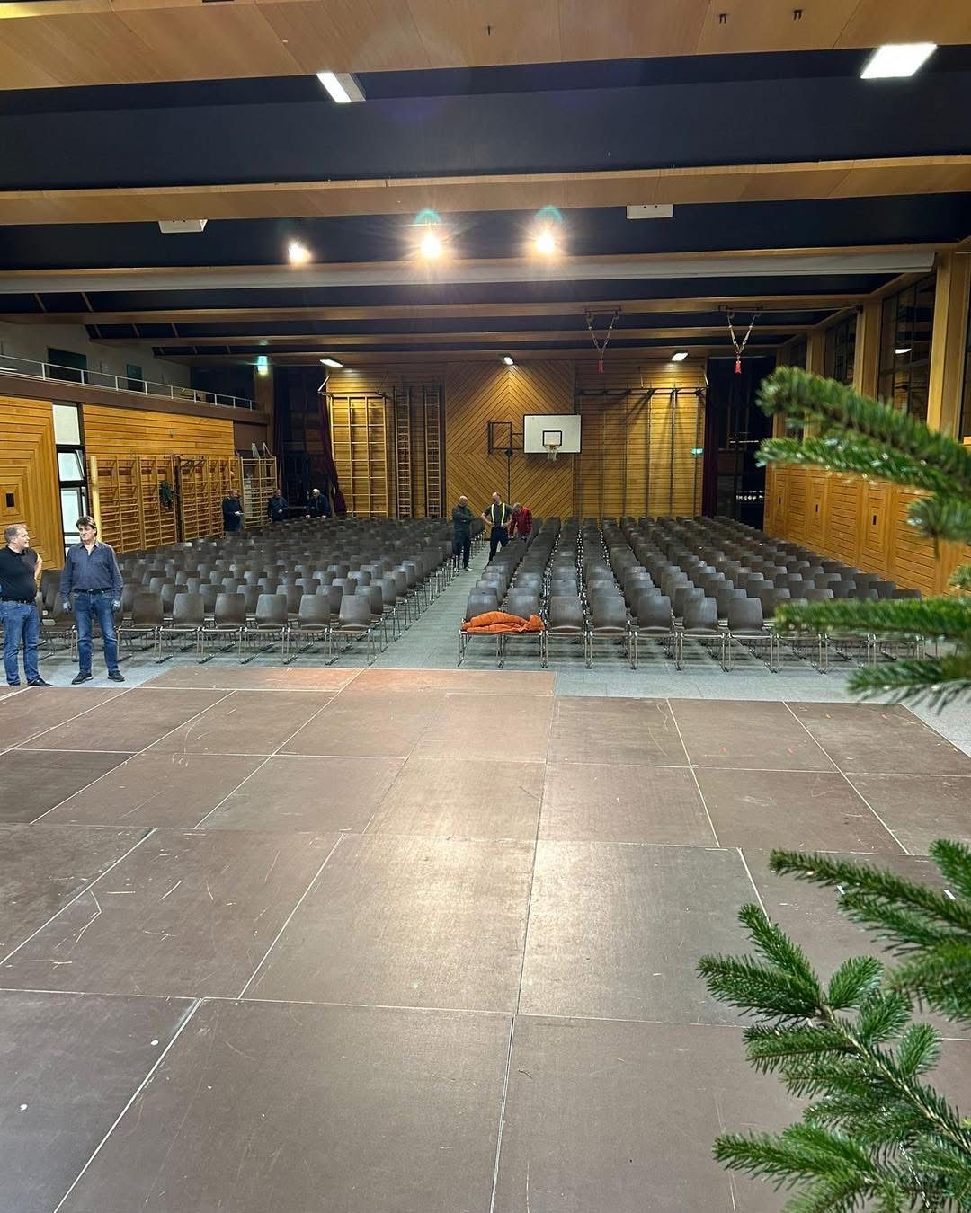 Ein leerer Saal mit Holzwänden und einem Basketballkorb. Mehrere Menschen gehen und stehen.