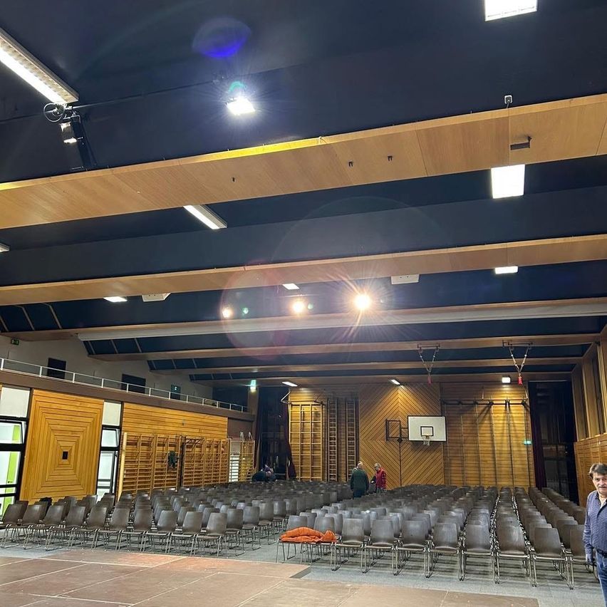 Ein leerer Saal mit Holzwänden, einem Basketballkorb und einer Bühne. Mehrere Personen stehen in der Nähe der Rückseite. Der Raum hat eine blaue Decke und einen Boden und viele Lichter.