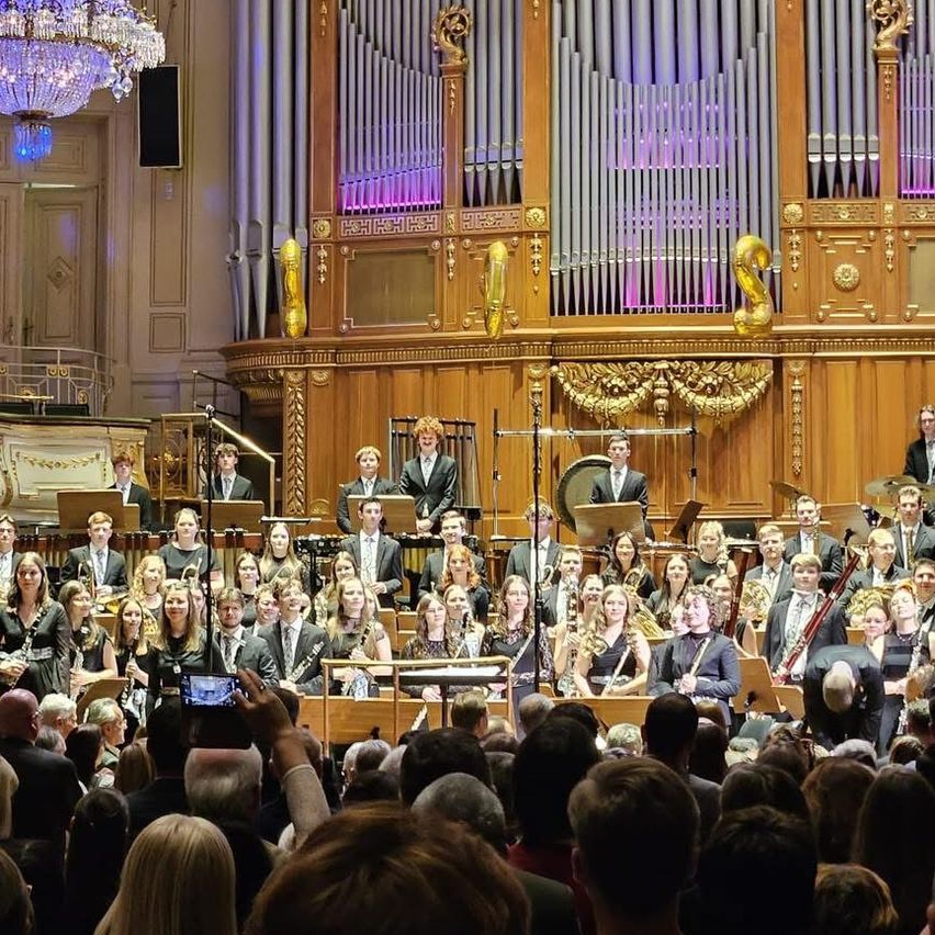 Ein großes Orchester und Chor treten auf einer Bühne auf, mit einer großen Orgel dahinter. Das Publikum sitzt davor, und einige machen Fotos. Die Darsteller sind formell gekleidet.