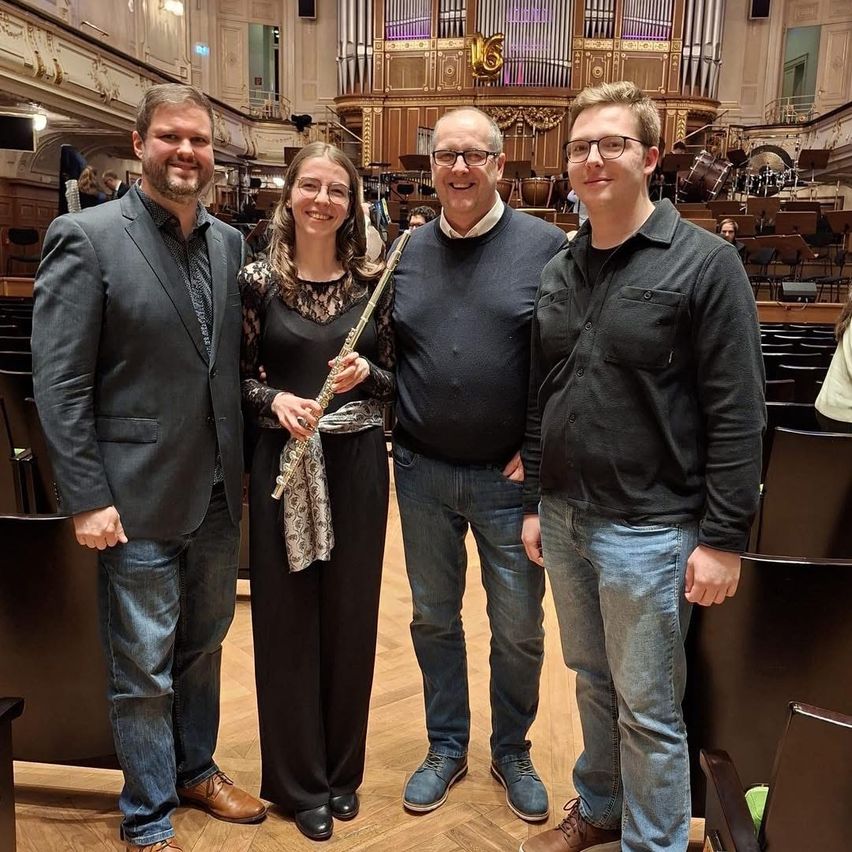 Vier Personen stehen in einem Auditorium, drei Männer in Freizeitkleidung und eine Frau mit einer Flöte. Sie lächeln und stehen nebeneinander. Dahinter ist ein Orchester zu sehen, Musiker sitzen an ihren Instrumenten.
