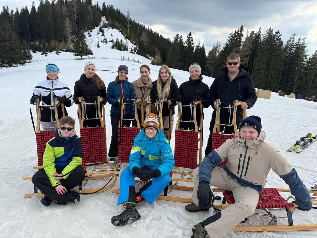 A group of people in winter clothes are posing for a photo on a snowy mountain. They are sitting on sleds, wearing hats and sunglasses. The background features trees and a hill.