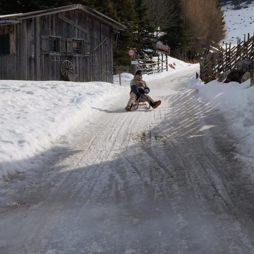 A person is sliding down a snowy road on a sled, with a wooden house and trees in the background.