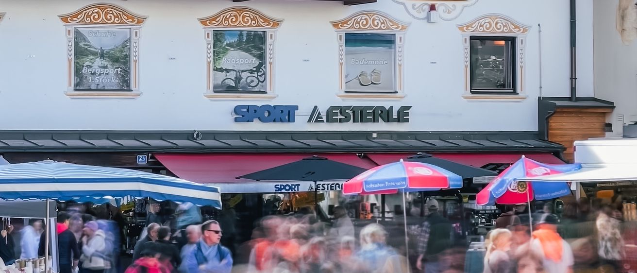 Ein geschäftiger Markt vor einem Gebäude mit Balkon, das mit einem roten Vordach und mehreren bunten Regenschirmen ausgestattet ist. Das Gebäude trägt ein Schild mit der Aufschrift 'SPORT & ESTERLE'. Menschen gehen herum, einige von ihnen tragen Sonnenbrillen.