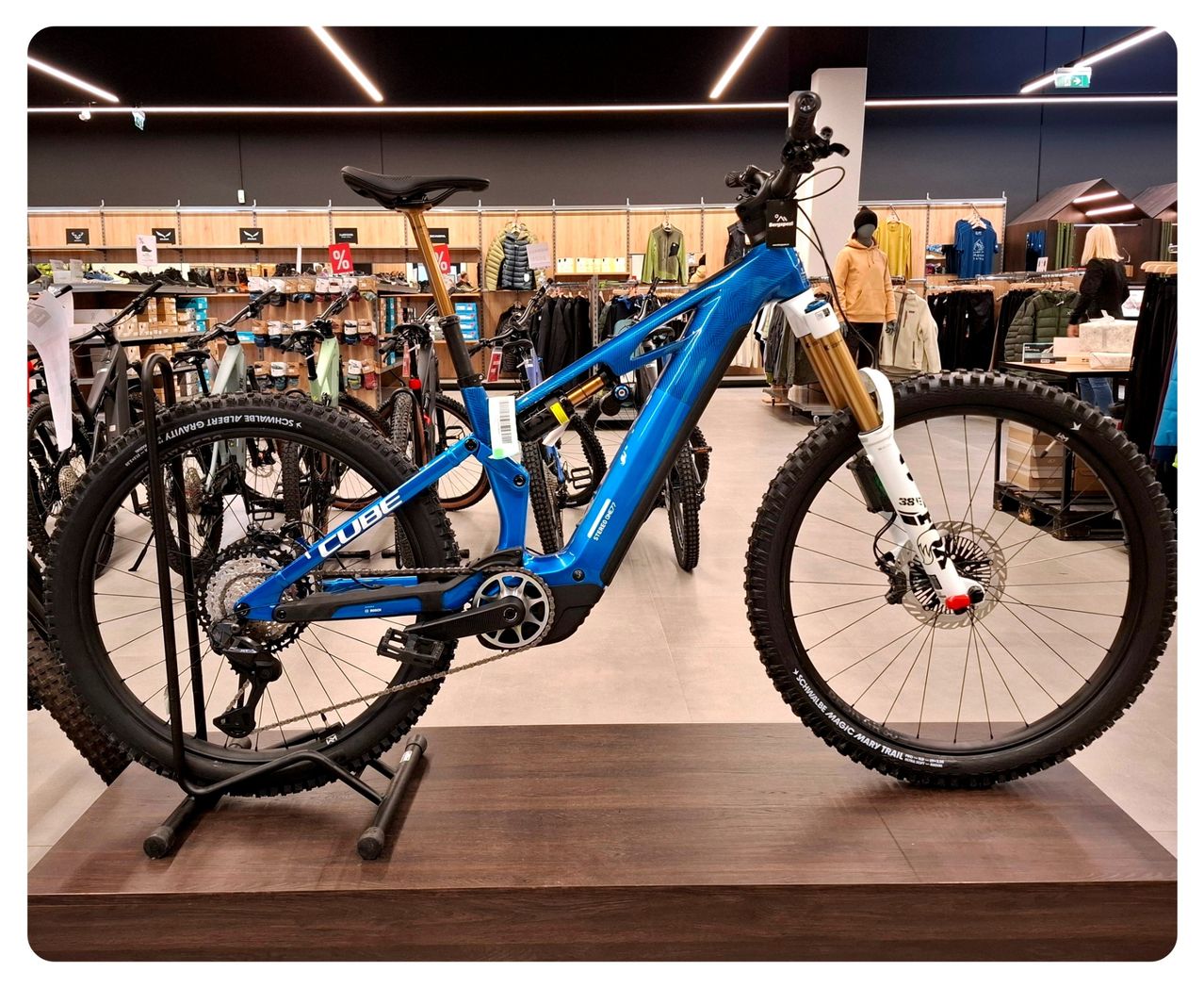 Ein blaues Mountainbike wird auf einer Holzplattform in einem Geschäft ausgestellt, umgeben von anderen Fahrrädern und Kleidung.