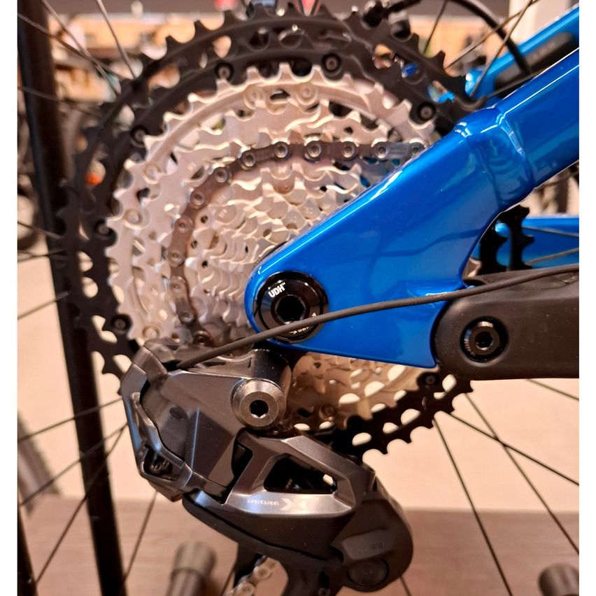 Nahaufnahme des Hinterderaillerleurs und Kettenblatts eines blauen Fahrrads. Der Derailleur ist metallisch und das Kettenblatt hat silberne und schwarze Zähne. Ein Draht verbindet den Derailleur mit dem Rahmen des Fahrrads.