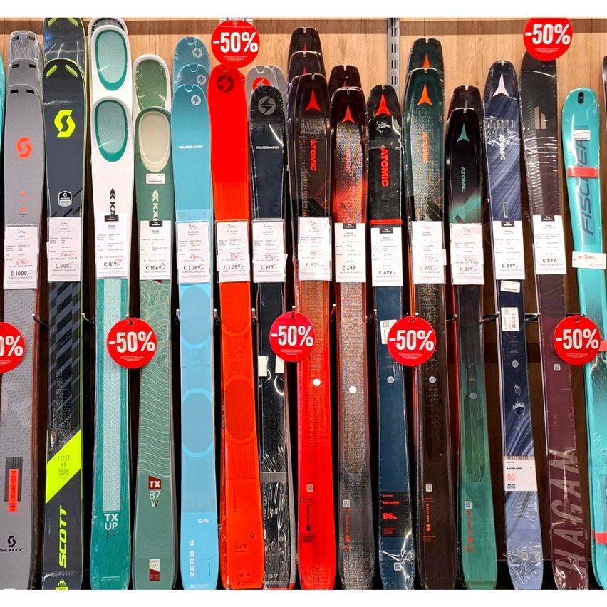 Eine Reihe von Skiern mit verschiedenen Designs und Farben ist ausgestellt und mit 50% Rabattetiketten versehen. Die Skier sind ordentlich angeordnet und präsentieren ihre unterschiedlichen Merkmale und Rabatte.
