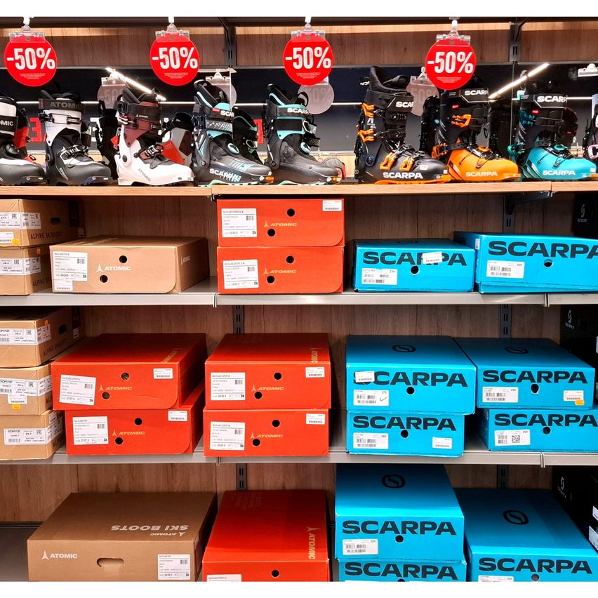 Ein Ladenregal zeigt verschiedene Snowboard-Schuhe mit erheblichen Rabatten. Kisten von Atomic-Snowboard-Schuhen und Scarpa-Snowboard-Schuhen sind in Reihen angeordnet. Rote und blaue Kisten sind gestapelt, einige mit Etiketten, und mehrere Schuhe sind am Regal montiert.