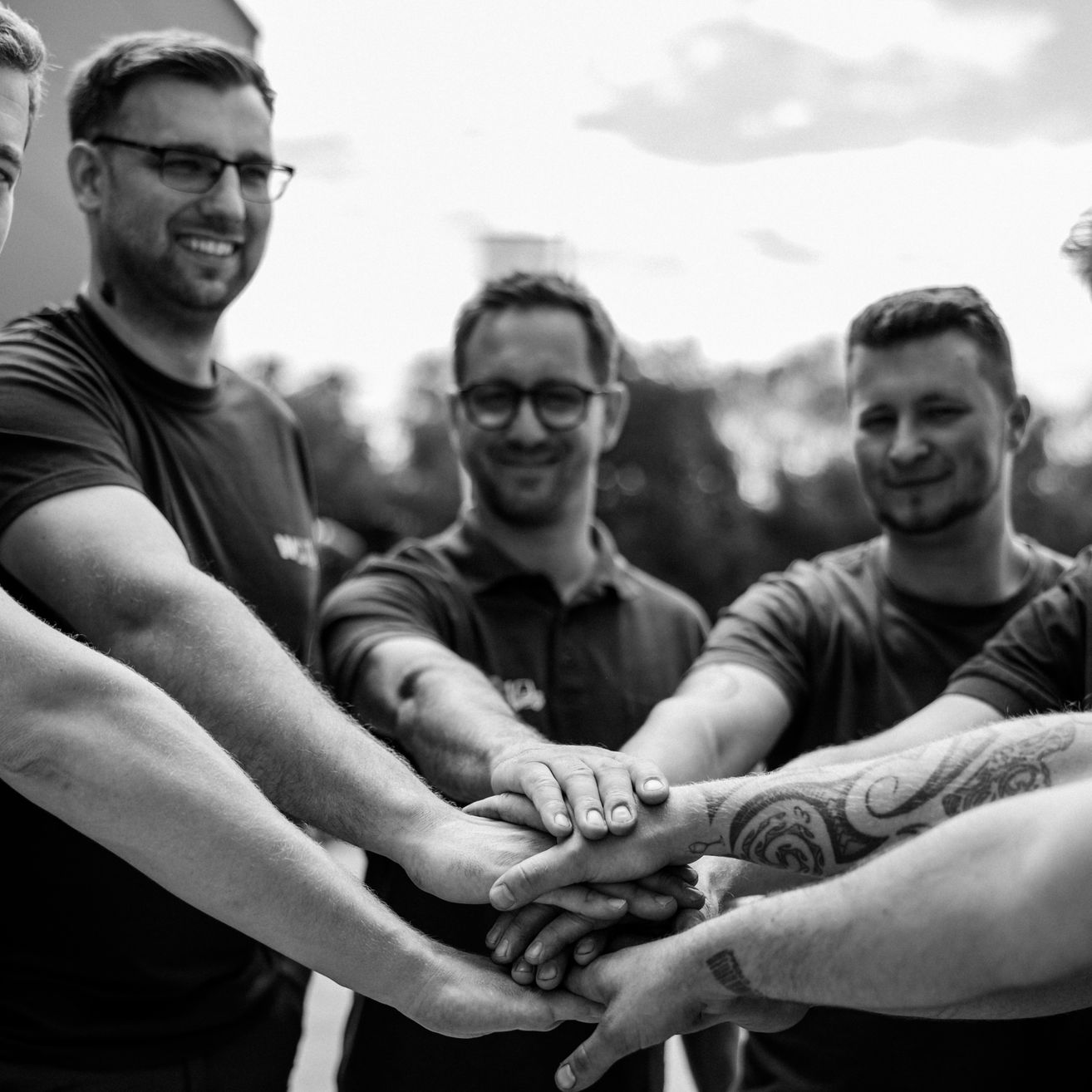 Fünf Männer in schwarzer und weißer Kleidung, möglicherweise bei einer Teambuilding-Aktivität, haben ihre Hände in der Mitte aufeinandergestapelt. Sie lächeln und stehen nebeneinander.