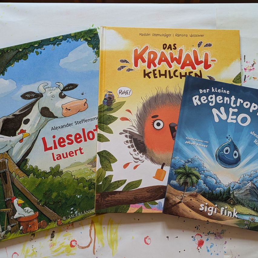 Drei Kinderbücher sind ausgestellt. 'Lieselo lauert' zeigt eine Kuh. 'Das Krawall-Kehlchen' stellt einen rotköpfigen Vogel dar. 'Der kleine Regentrop NEO' zeigt einen Regentropfen.