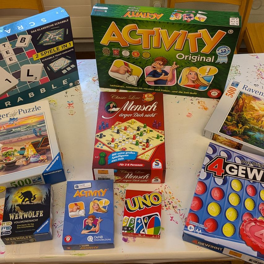 Auf einem Tisch sind verschiedene Brettspiele zu sehen. Dazu gehören Scrabble, Mensch Arger Dich Nicht, UNO und 4 Gewinnt. Jedes Spiel hat ein eigenes Design und Thema auf seiner Schachtel.