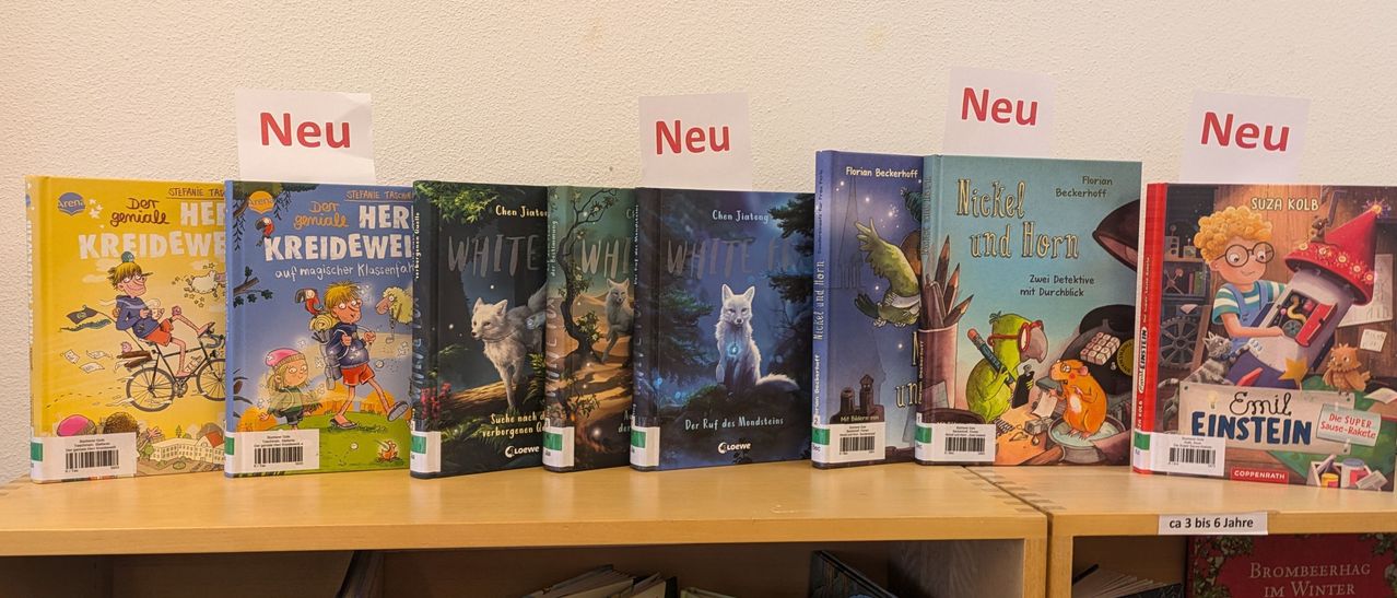 A row of books on a shelf includes five books with a white wolf on the cover. Two signs in red say 'Neu'. The last book has a bird and pencils on the cover.