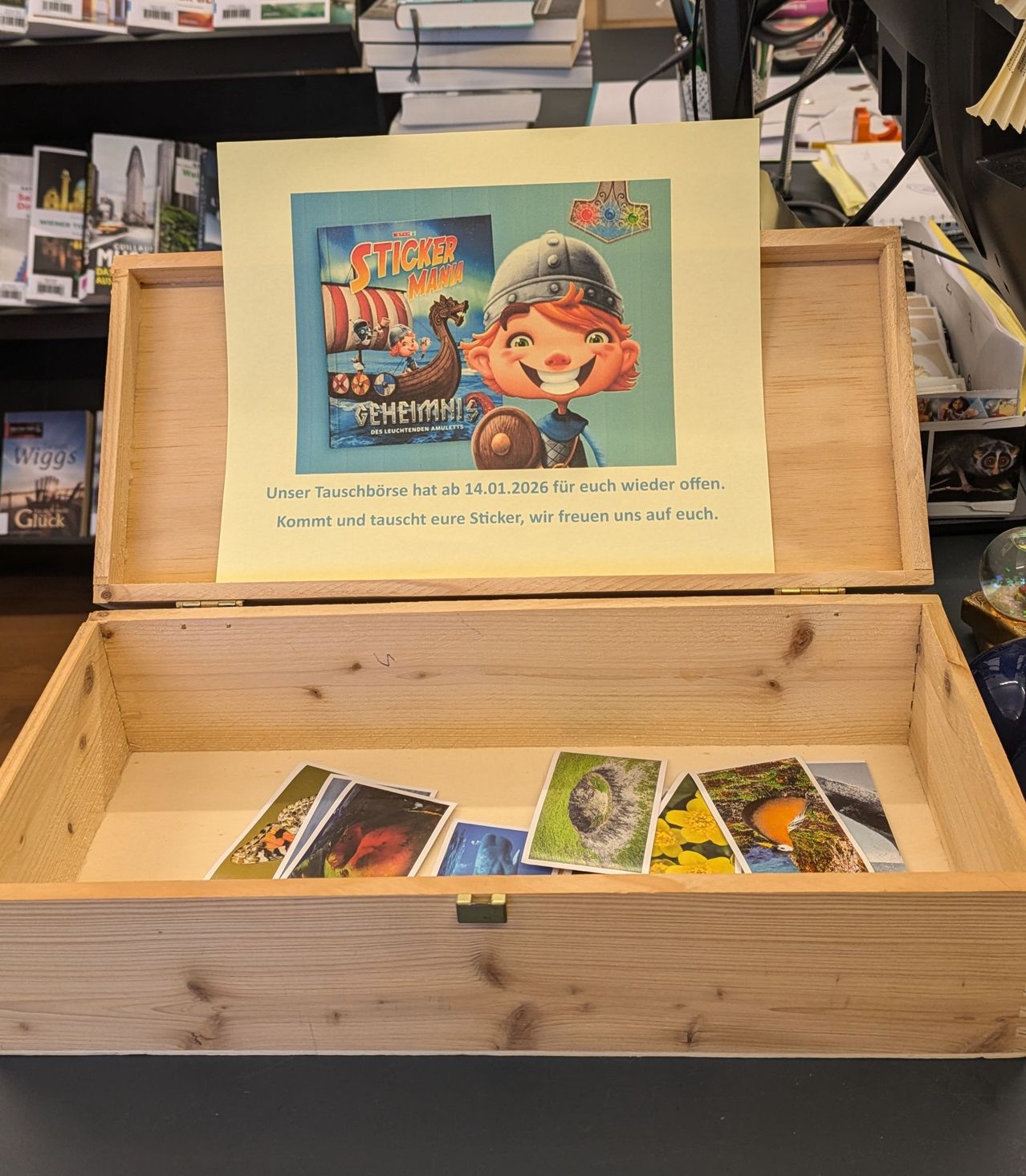 A wooden box with several cards inside. A sign on the box reads, 'Exchange your stickers until January 14, 2026. Come and trade your stickers, we look forward to seeing you.'