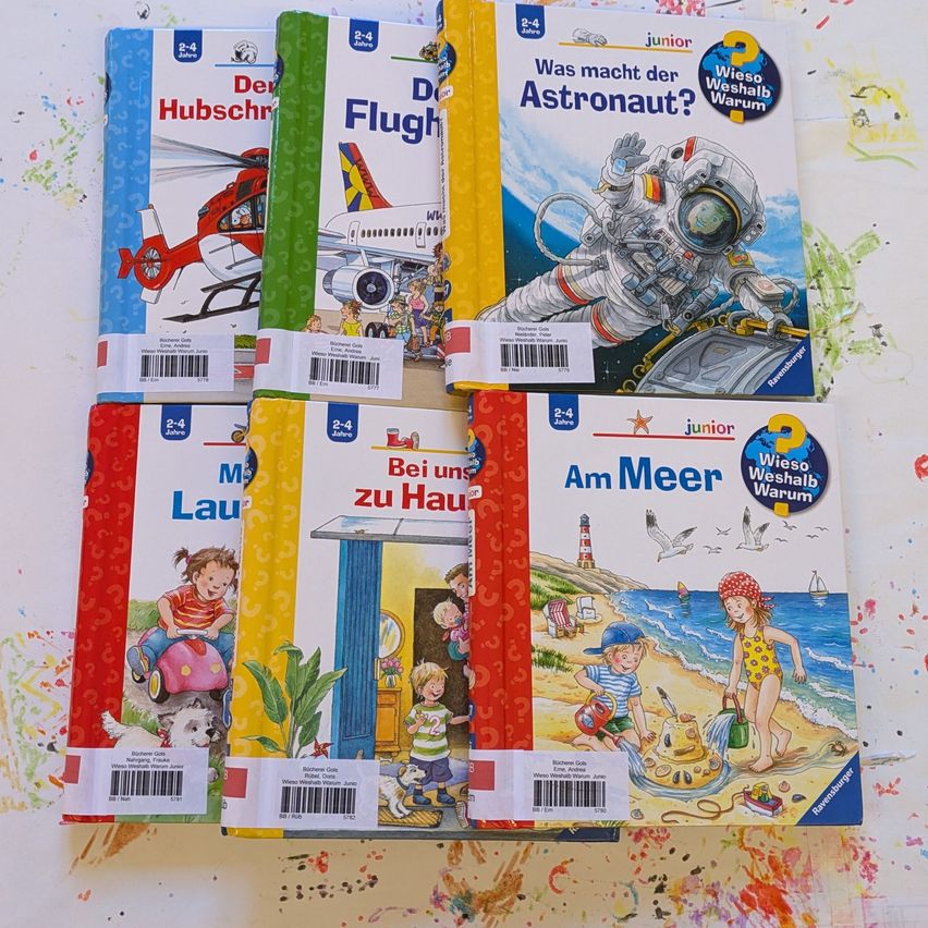 Eine Sammlung von sechs Kinderbüchern, die auf einer weißen Oberfläche ausgestellt sind. Jedes Buch zeigt farbenfrohe Illustrationen und Text in Deutsch. Titel beinhalten 'Der Hubschrauber', 'Der Flugzeug', 'Was macht der Astronaut?', 'Bei uns zu Haus', 'Mauer Lauf', und 'Am Meer'.