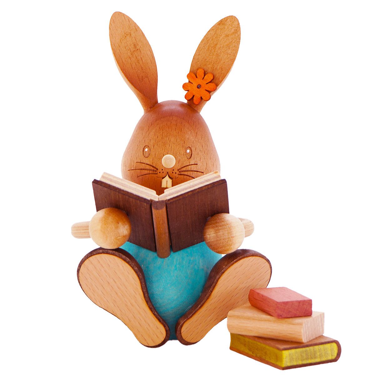 Eine Holzfigur eines Kaninchens sitzt auf einem weißen Hintergrund und liest ein Buch. Das Kaninchen trägt ein blaues Shirt und hat eine Blume an seinem Ohr. Daneben sind gestapelte Holzbücher.