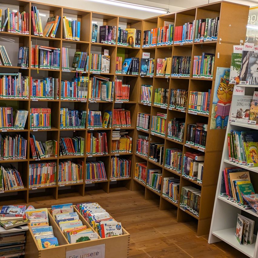 Eine Bibliothek mit Holzregalen voller bunter Bücher, einige sind als Neu gekennzeichnet. Ein Schild sagt 'Fur unsere'.