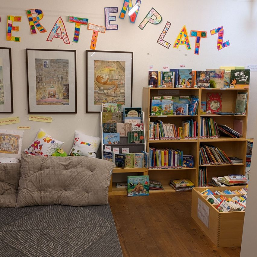 Eine Leseecke mit Kissen und einer Couch, umgeben von Bücherregalen voller Kinderbücher, und farbenfrohe Worte an der Wand, die 'TERATEMPLATZ' sagen.