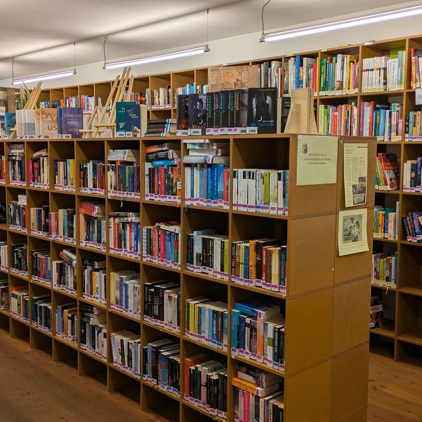 Rows of bookshelves filled with various books in a library. Wooden shelves and a yellow sign on one of the shelves. The library has a wooden floor and white ceiling lights.