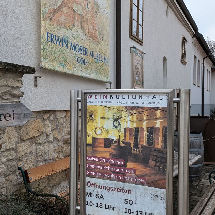 A wine culture house displays opening hours on a sign in front of a stone wall. A wooden bench is beside it. A large banner with Erwin Moser Museum and Gols is mounted on the wall. Two barrels are on the right side.