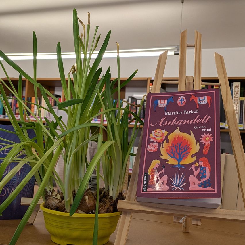 A book titled 'Ankandelt' by Martina Parker is displayed on an easel next to a green plant in a yellow pot, inside a library with bookshelves in the background.