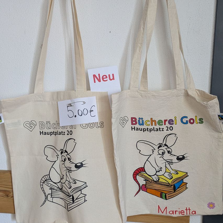 Zwei beige Stofftaschen mit einer Maus, die Bücher liest. Eine hat den Text 'Bücherei Gols Hauptplatz 20' und die andere hat 'Bücherei Gols Hauptplatz 20'. Ein Preisschild von 5,00€ ist an der ersten Tasche befestigt.