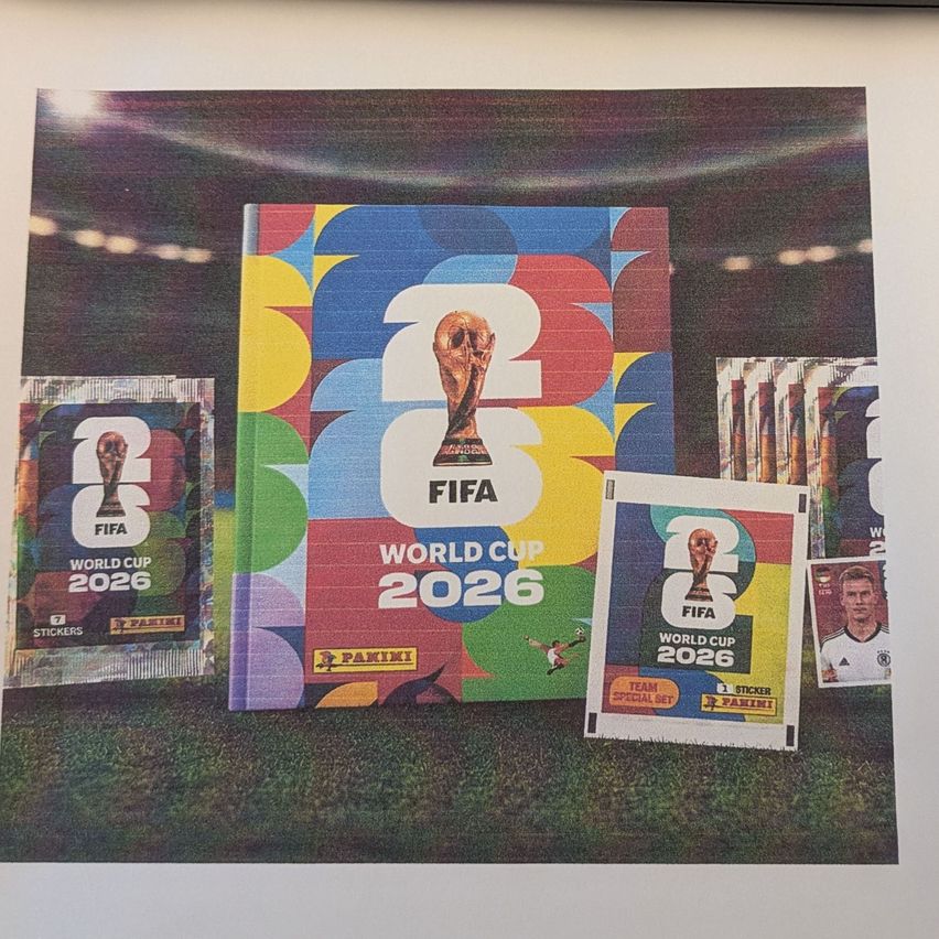 Eine Sammlung von FIFA Weltmeisterschaft 2026 Merchandise, einschließlich eines Buches, Aufklebern und einer Spielerkarte, die auf einem grünen Feldhintergrund ausgestellt sind.