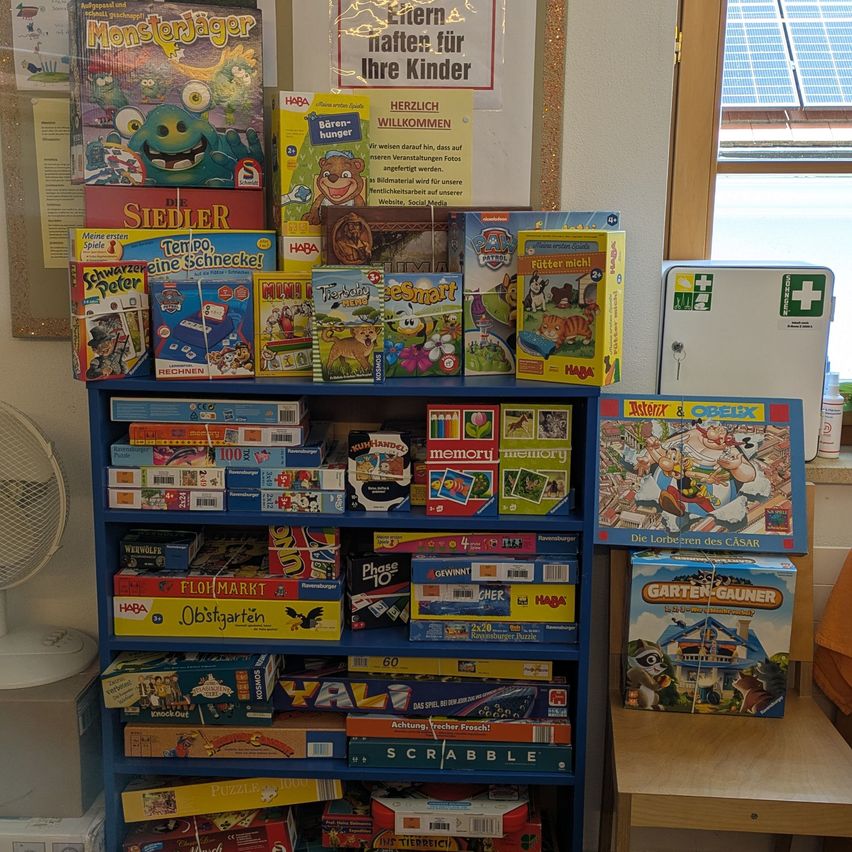 Ein blaues Bücherregal gefüllt mit verschiedenen Brettspielen, darunter Titel wie 'Monsterjäger', 'Die Siedler' und 'Tempo, eine Schnecke!'. Ein Schild an der Wand steht 'Eitern naften für Ihre Kinder'. Ein Ventilator steht links vom Regal.