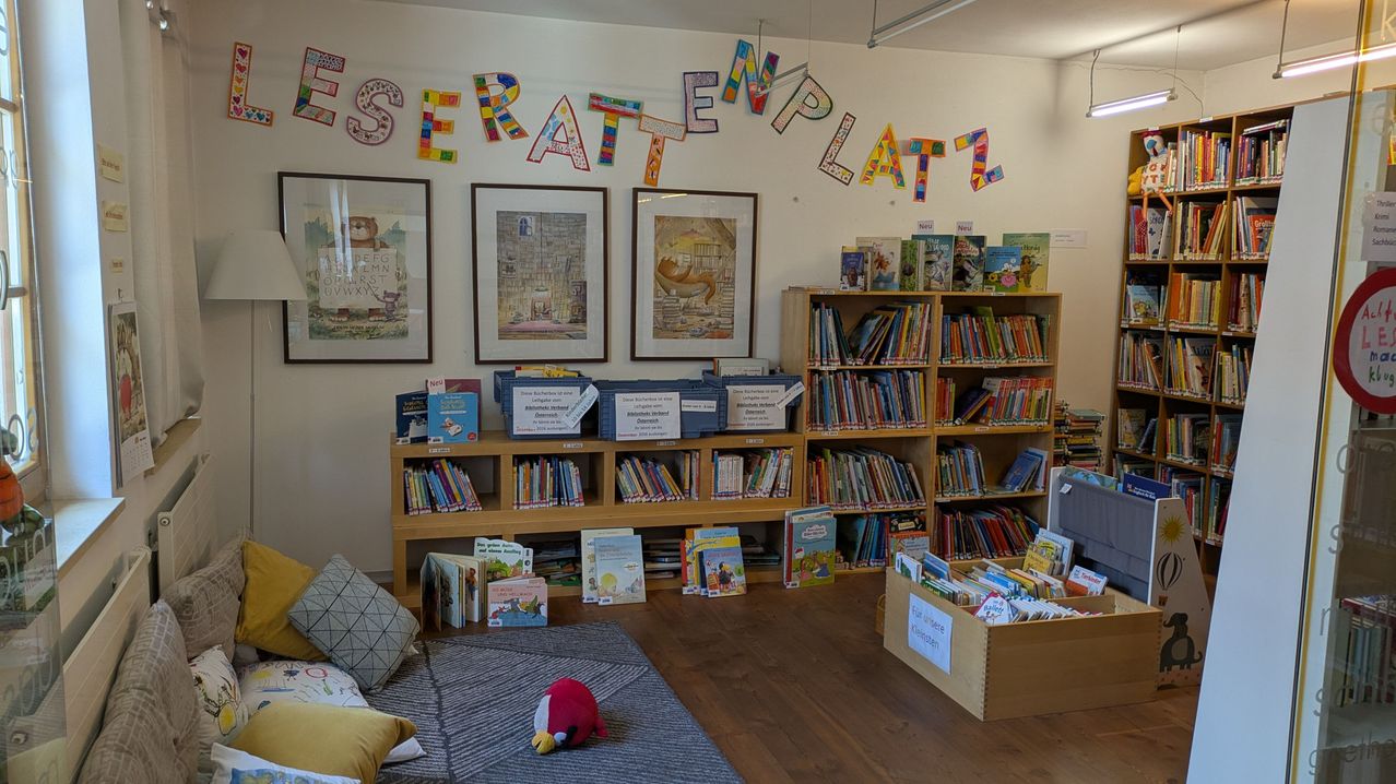Eine gemütliche Leseecke in einer Bibliothek mit bunten Buchstaben am Wand, einem hölzernen Bücherregal voller Bücher und einem roten Angry Birds Plüschtier auf dem Boden.