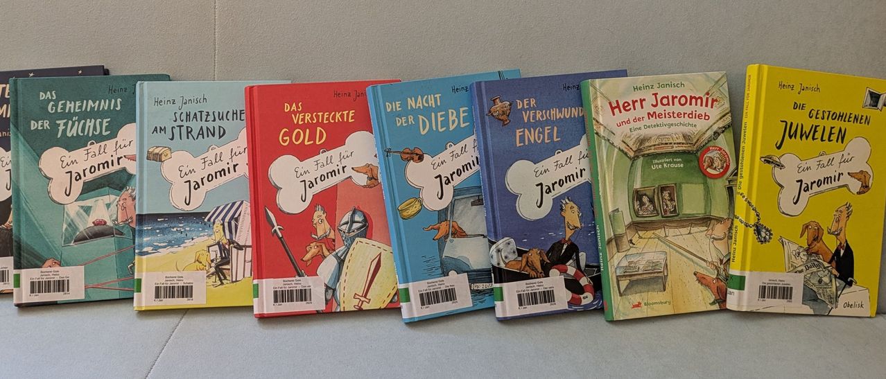 Fünf Bücher mit den Titeln 'Das Versteckte Gold', 'Die Nacht der Diebe', 'Der Verschwunden Engel', 'Ein Fall für Jaromir' und 'Heinz und Jaromir' sind auf einem Tisch aufgereiht. Jedes Buch hat ein eigenes Cover-Design mit Illustrationen eines Hundes, eines Ritters, eines Diebes, eines Engels und eines Mannes in einem Boot.