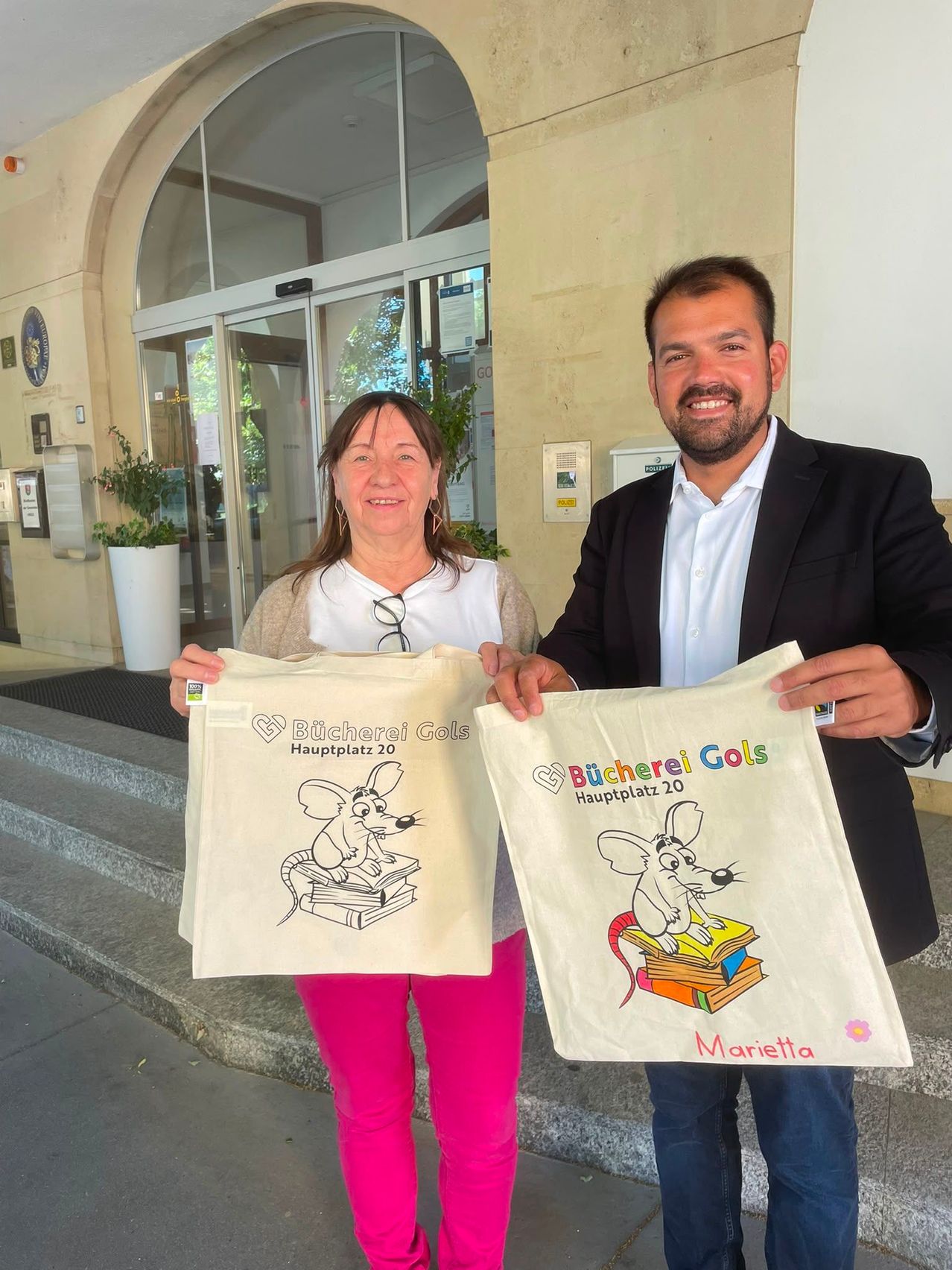 A man and a woman are standing in front of a building, holding bags with a mouse illustration and the text 'Bucherei Gols Hauptplatz 20'. The woman is wearing glasses and pink pants, while the man is in a suit.