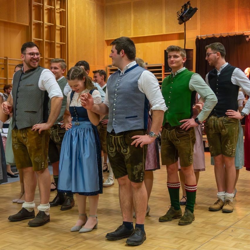Mehrere junge Männer und Frauen in traditioneller bayerischer Kleidung stehen in einer Reihe, möglicherweise in einer Tanzhalle, und sehen sich gegenseitig an.