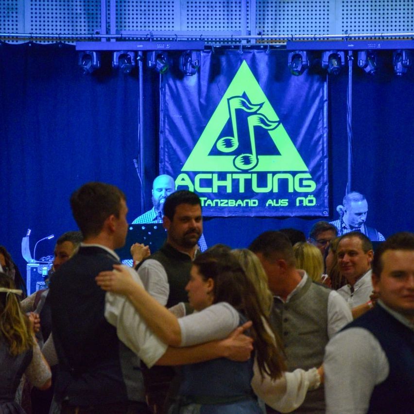 Eine Gruppe von Menschen tanzt bei einer Veranstaltung mit einem blauen Hintergrund und einem grünen Musiknoten-Schild. Eine Person im Hintergrund scheint aufzutreten.