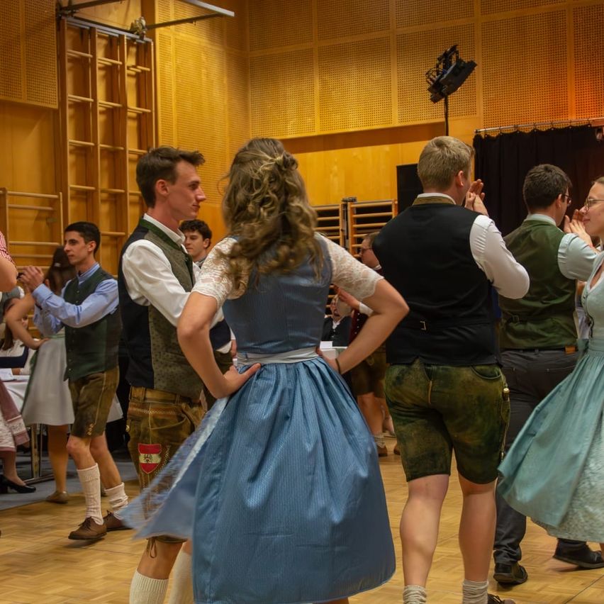 Eine Gruppe von Menschen in traditioneller Kleidung tanzt gemeinsam in einem Saal. Eine Frau in der Mitte hat die Hände auf der Taille. Dahinter tanzen andere, und einige stehen an der Wand.