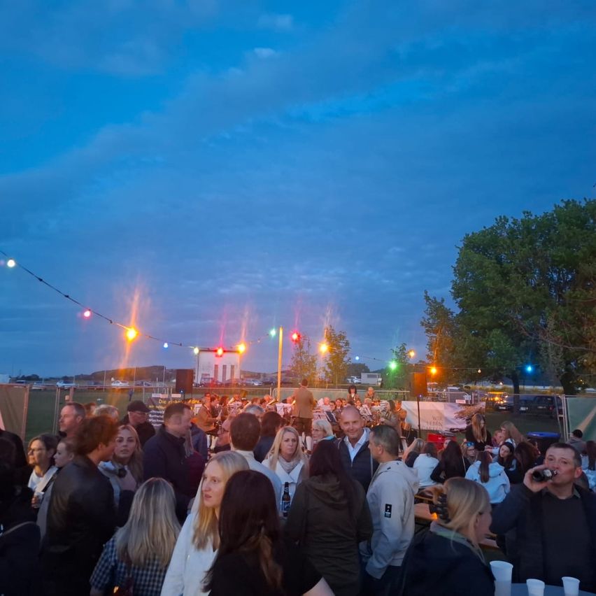 A large crowd of people, likely at an outdoor event, are gathered under a blue sky with some clouds. The area is illuminated by string lights and various colored lights. Trees and a building are visible in the background.