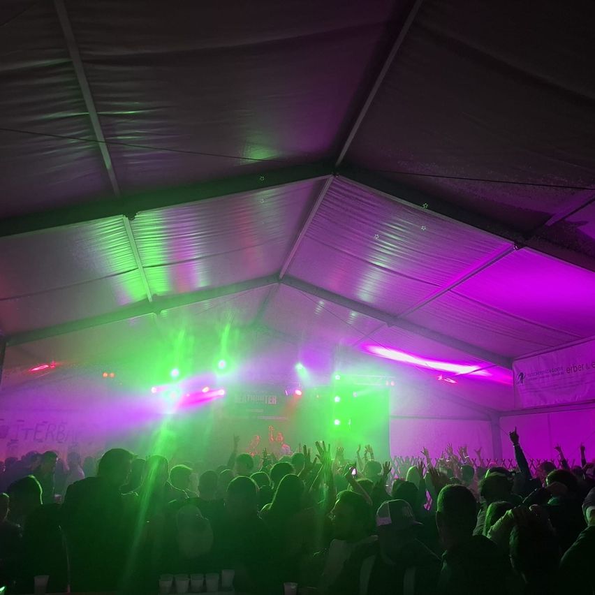 A crowd under a tent at a concert, with purple and green lights illuminating the scene. Many people are raising their hands, and cups are visible on the table in front.