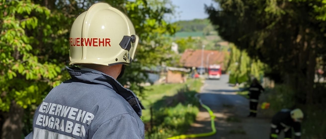 Ein Feuerwehrmann mit der Aufschrift 'Feuerwehr Eugengraben' auf dem Rücken schaut auf einen gelben Schlauch, der auf dem Boden liegt. Ein anderer Feuerwehrmann repariert den Schlauch.
