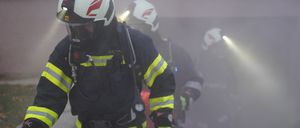 Feuerwehrleute in voller Ausrüstung gehen durch einen rauchigen Bereich, mit Helmen und Masken. Ein Feuerwehrmann führt, ein anderer folgt dicht dahinter. Ein dritter Feuerwehrmann ist im Hintergrund, ebenfalls in Schutzausrüstung.