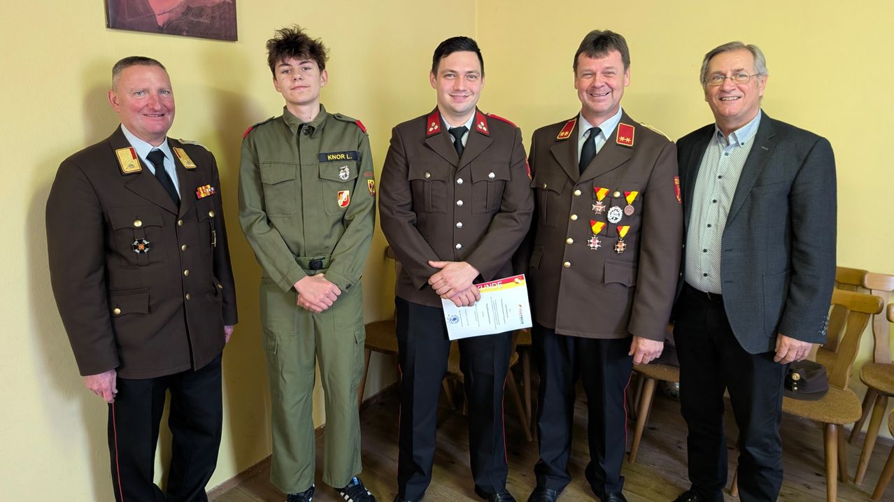 Drei Männer in Militäruniformen stehen nebeneinander, einer hält ein Zertifikat in der Hand, lächeln und posieren für ein Foto in einem Raum mit gelber Wand.