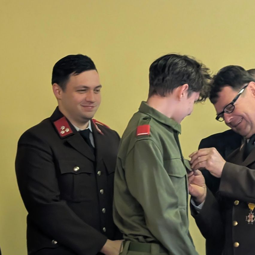 Drei Männer in Militäruniformen stehen eng beieinander. Einer hält eine Medaille, ein anderer lächelt und der dritte scheint die Medaille an der Uniform des zweiten Mannes zu befestigen.