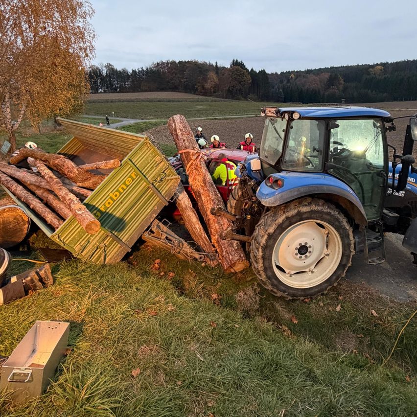 Ein blauer Traktor liegt mit umgestürzten Baumstämmen und einem Anhänger auf einem Grasfeld. Rettungskräfte in Helmen umgeben das Fahrzeug.