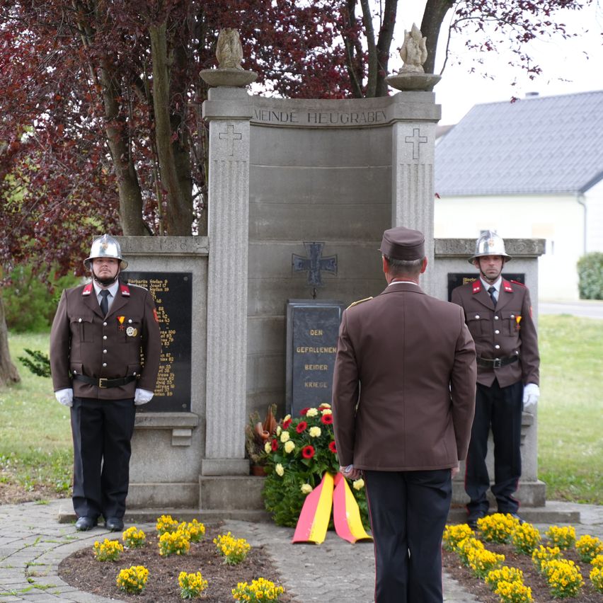 Drei uniformierte Männer stehen an einem Denkmal mit einem Kreuz, Blumen und einer Tafel mit deutschem Text. Der Mann vorne steht mit dem Rücken zur Kamera.