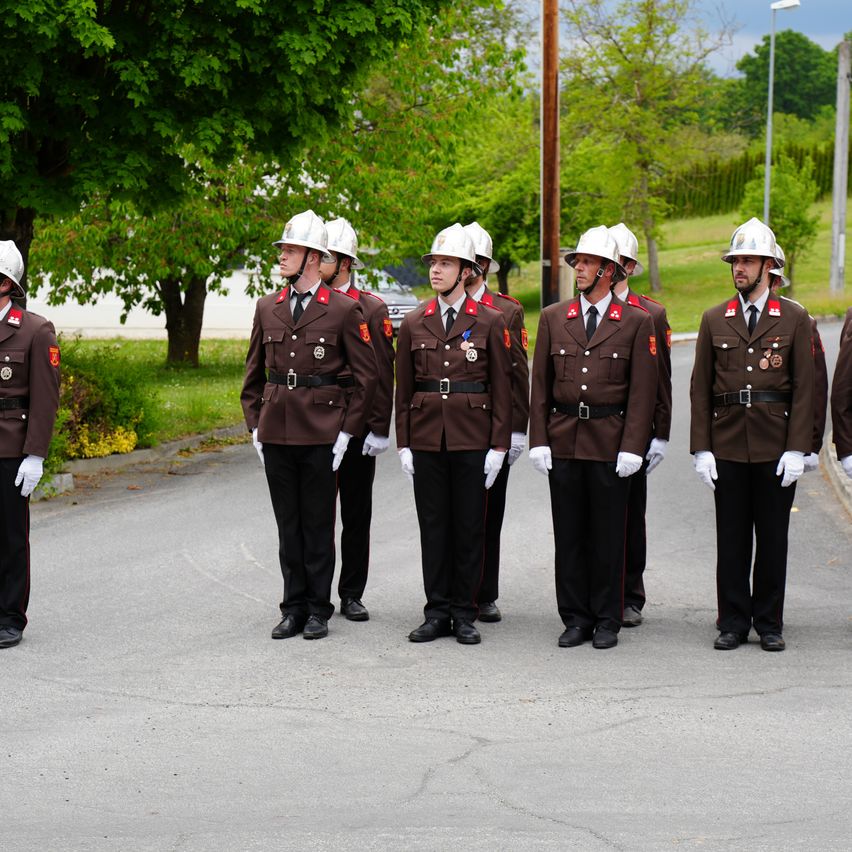 Sechs Personen in Militäruniformen stehen in einer Reihe, weiße Handschuhe und Helme tragend. Sie stehen auf einem gepflasterten Weg mit Grünflächen und Bäumen im Hintergrund.