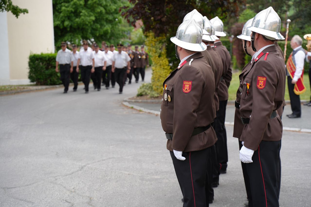 Eine Gruppe uniformierter Personen, wahrscheinlich Feuerwehrleute, stehen in einer Reihe mit Helmen und weißen Handschuhen. Eine weitere Gruppe marschiert hinter ihnen.