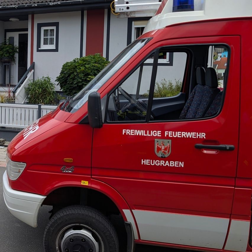 Ein rotes Fahrzeug der Freiwilligen Feuerwehr ist vor einem weißen Haus mit rotem Dach geparkt. Das Fahrzeug hat die Aufschrift Freiwillige Feuerwehr Heugraben an der Seite.